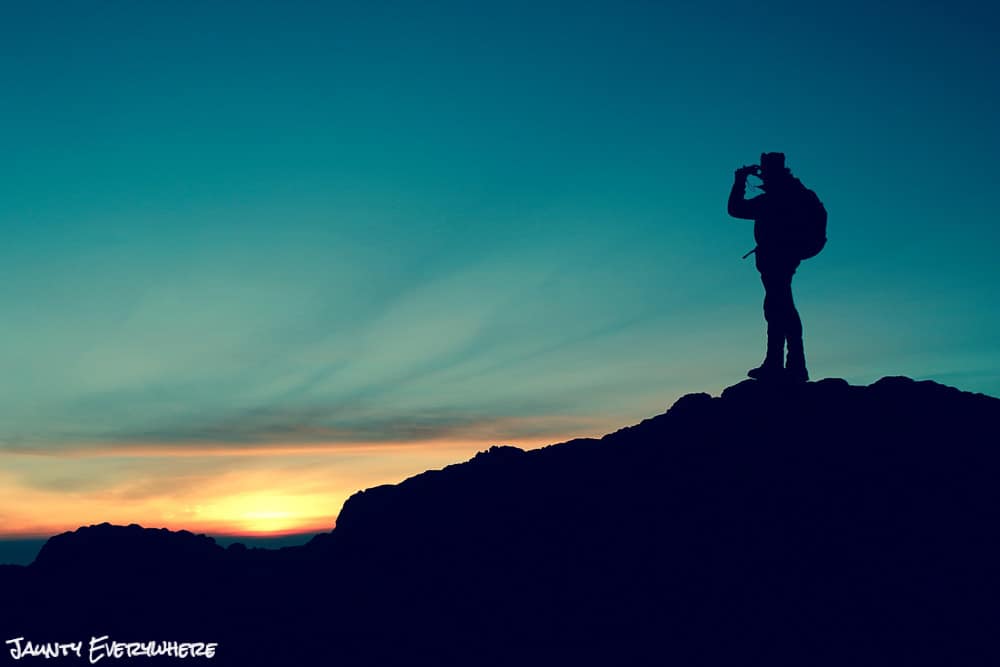 hiker standing on a mountain watching the sunrise through binoculars