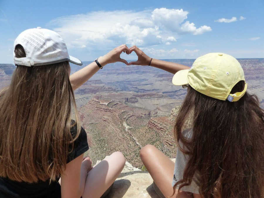 two young women sitting over the Grand Canyon, their hands are together with fingers forming the shape of a heart