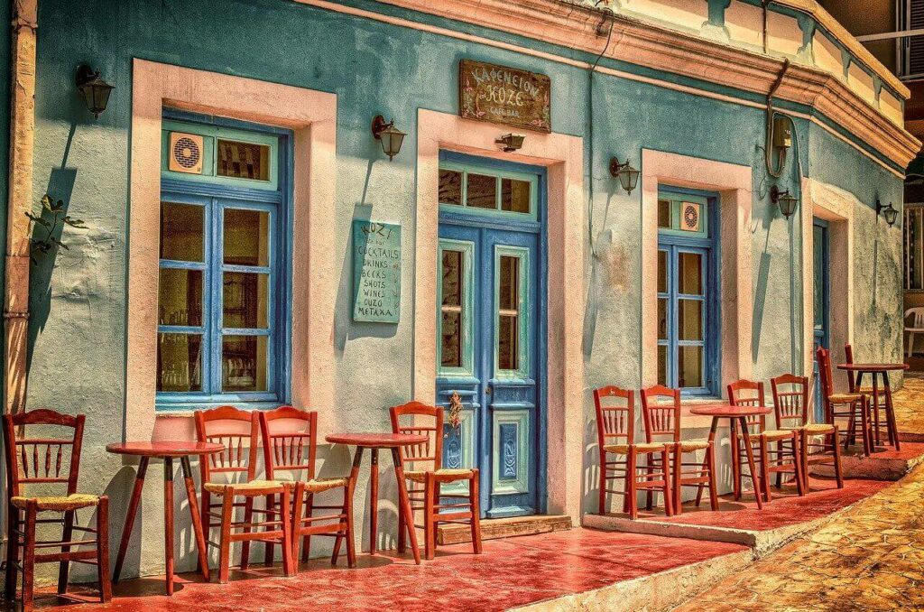 the front of a Greek cafe, turquoise stucco and red cafe chairs and tables line the front