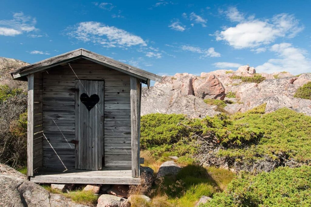 wooden outhouse with a heart shaped cut out in the door, set on a rocky outcropping 