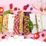 a variety of granola bars lined up artistically and sprinkled with oats and berries on a pink background