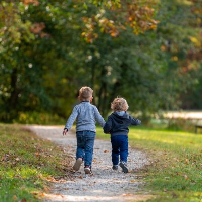 two small boys running down a path under large trees.