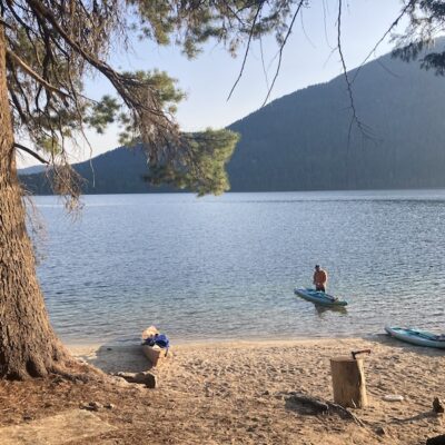A lone paddle boarding stands out in the water, working on his paddle board. The lake is blue and the sun is casting a warm glow over everything.